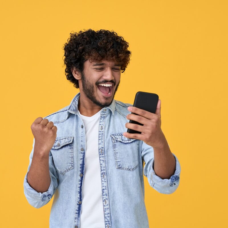 Excited happy young indian man winner feeling joy using smartphone winning lottery game, betting, getting cashback online gift in mobile app message holding cell phone isolated on yellow background.